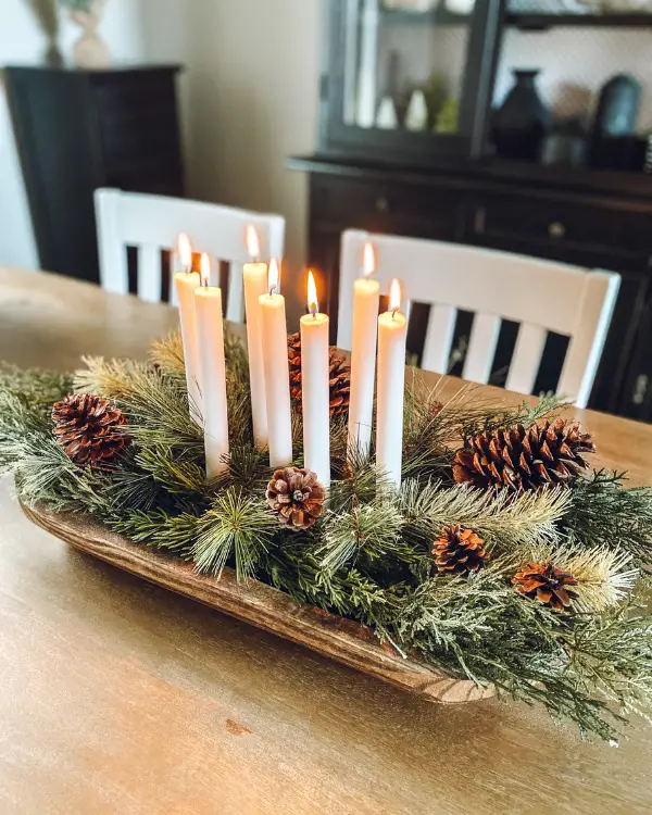 A Christmas centerpiece with lit candles, greenery, pinecones and a dough bowl